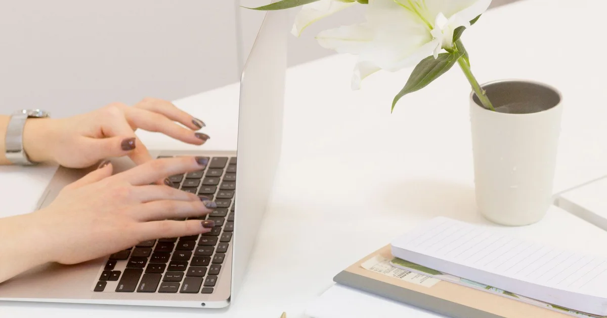 Hands typing at a bright desk with a laptop and office supplies.